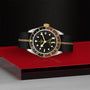 The TUDOR Black Bay GMT S&G luxury watch, inspired by Tudor divers’ models, showcases a black and gold face and a black strap with a gold stripe, displayed on red and gray surfaces against a textured black background.