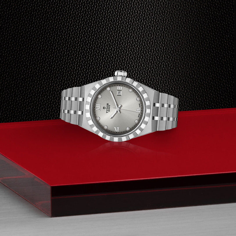 A TUDOR Royal silver wristwatch with a metal bracelet is displayed on a red and black textured surface.