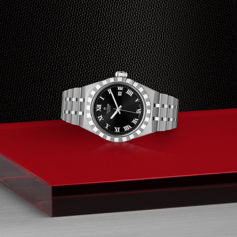 A TUDOR Royal automatic sport-chic watch with a black Roman numeral dial and two-tone bracelet is displayed on a red and gray platform against a textured black background.