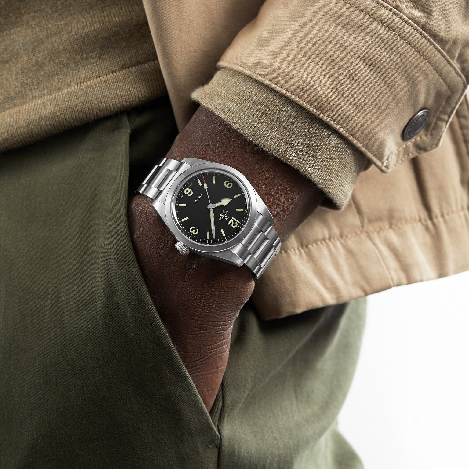 A person in a beige jacket and green pants has their hand in their pocket, showing a TUDOR Ranger watch with a black dial, Arabic numerals, and Super Luminova for enhanced visibility.