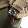 A person in a beige jacket and green pants has their hand in their pocket, showing a TUDOR Ranger watch with a black dial, Arabic numerals, and Super Luminova for enhanced visibility.