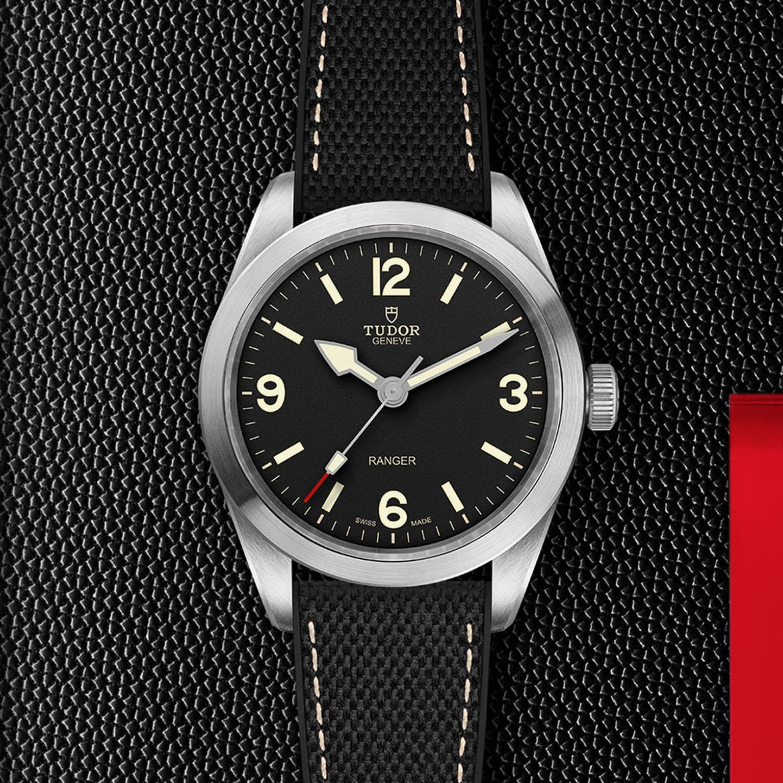 A TUDOR Ranger wristwatch featuring a black dial, beige Super Luminova numerals, and a black textured strap is showcased on a dark textured background.