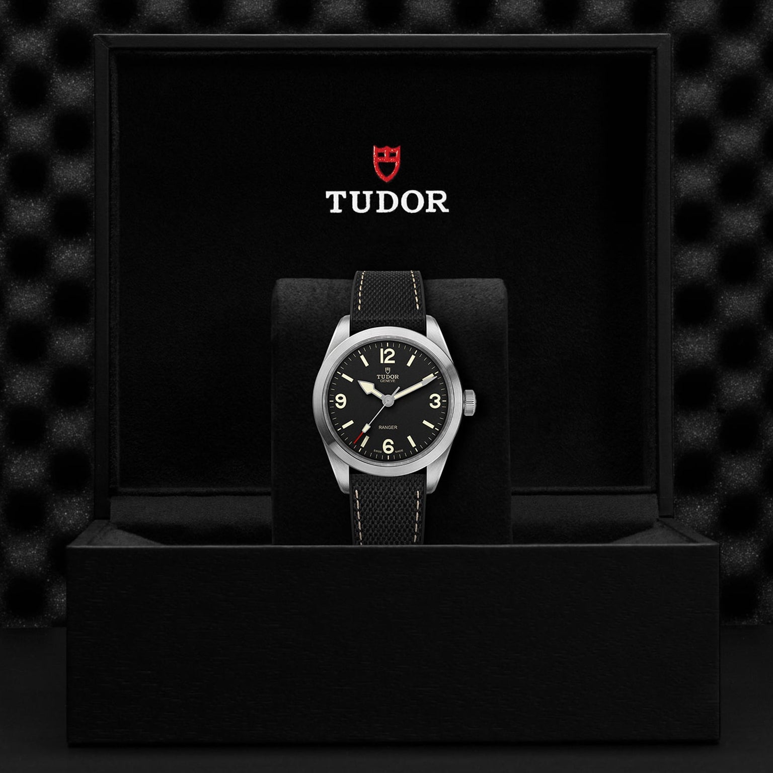A TUDOR Ranger wristwatch with a black dial and fabric strap, enhanced by Super Luminova accents, is displayed in an open black TUDOR presentation box.