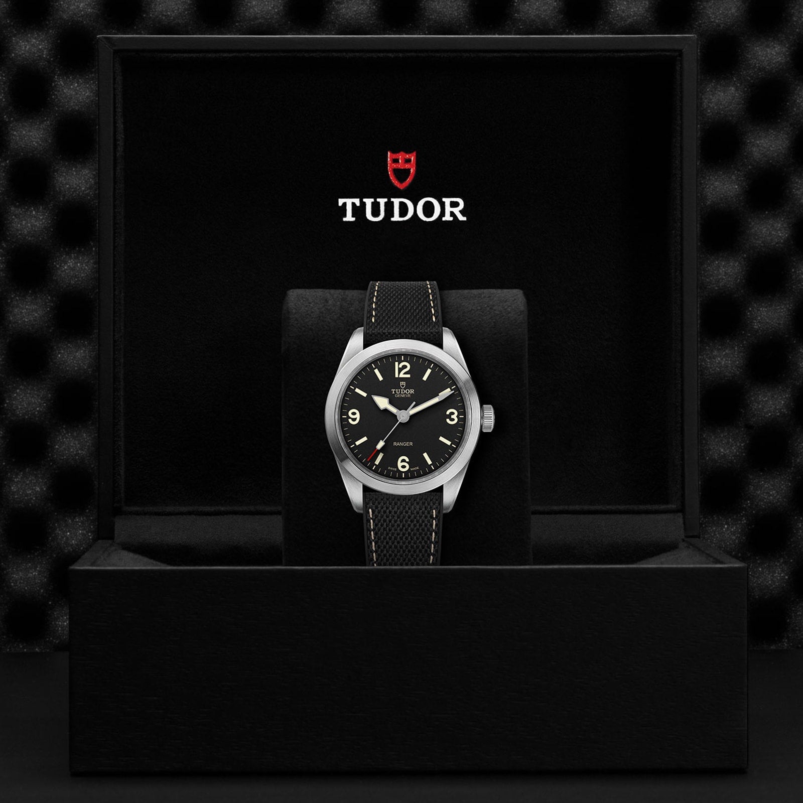 A TUDOR Ranger wristwatch with a black dial and fabric strap, enhanced by Super Luminova accents, is displayed in an open black TUDOR presentation box.