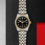 The TUDOR Black Bay One 36 S&G features a black dial with yellow gold hour markers and a two-tone gold and silver bracelet, elegantly displayed on a textured black surface.