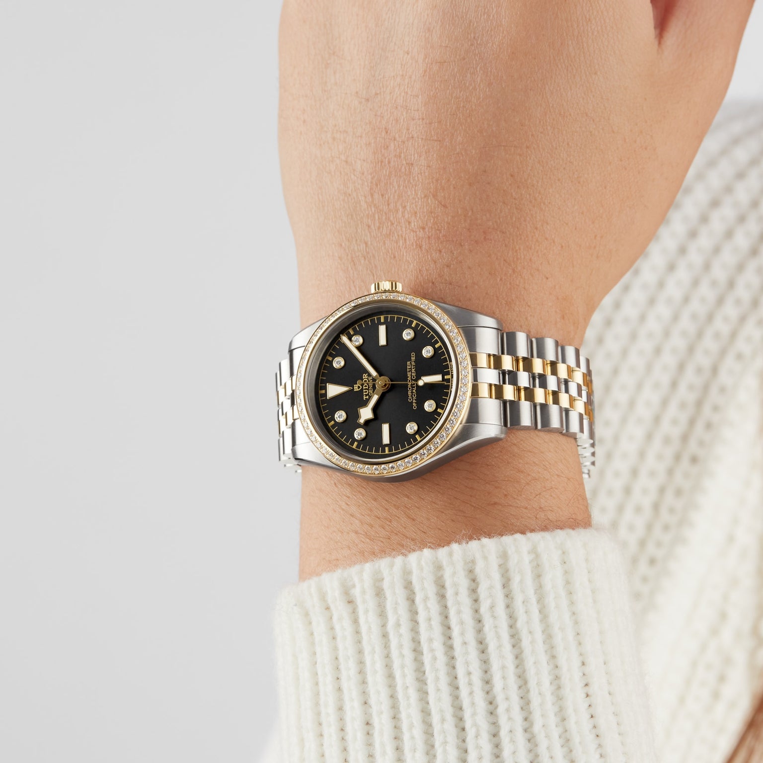 A person wears a TUDOR Black Bay One 36 S&G wristwatch featuring a black dial and yellow gold accents; their hand rests against a neutral background, with part of a white sweater visible.