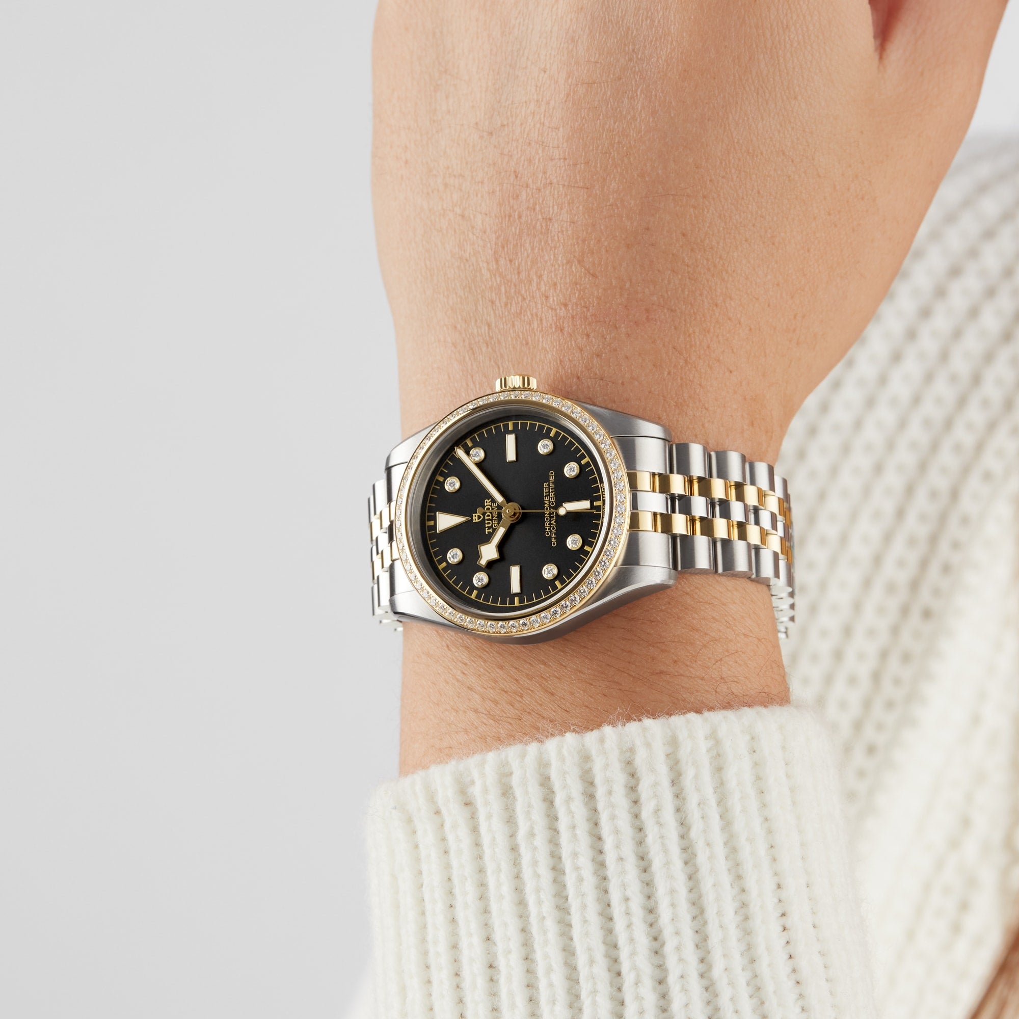 A person wears a TUDOR Black Bay One 36 S&G wristwatch featuring a black dial and yellow gold accents; their hand rests against a neutral background, with part of a white sweater visible.