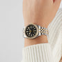 A person wears a TUDOR Black Bay One 36 S&G wristwatch featuring a black dial and yellow gold accents; their hand rests against a neutral background, with part of a white sweater visible.