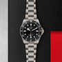 The TUDOR Pelagos 39, a stainless steel wristwatch with a black dial and bezel, is displayed on a textured black surface with metallic and red accents—an outstanding professional dive watch for mechanical watch enthusiasts.