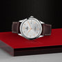 The TUDOR 1926, a classic silver wristwatch with a brown leather strap, features a mechanical date display and silver-gold hour markers. It rests elegantly on a red and black geometric surface.