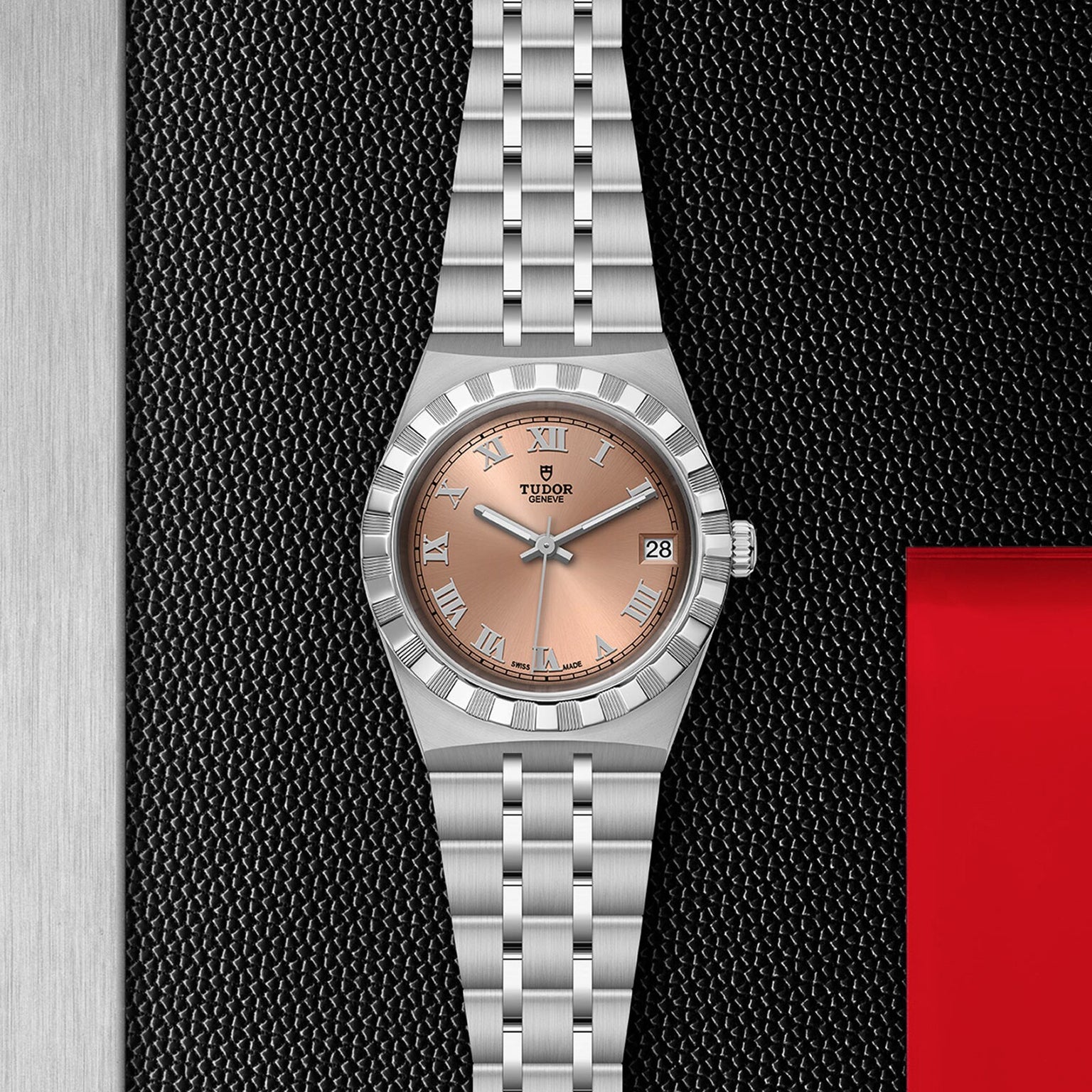 The TUDOR Royal is a chic stainless steel wristwatch with a salmon dial, Roman numerals, date display, and silver link bracelet, shown on a black and red textured background.