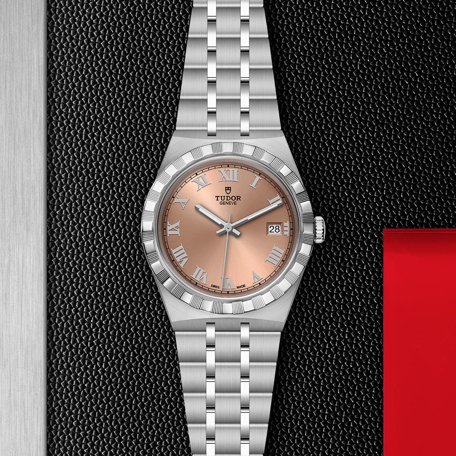 A TUDOR Royal wristwatch with a salmon dial, Roman numerals, date display, and stainless steel bracelet showcases sport chic style and automatic movement as it rests on a textured black surface.