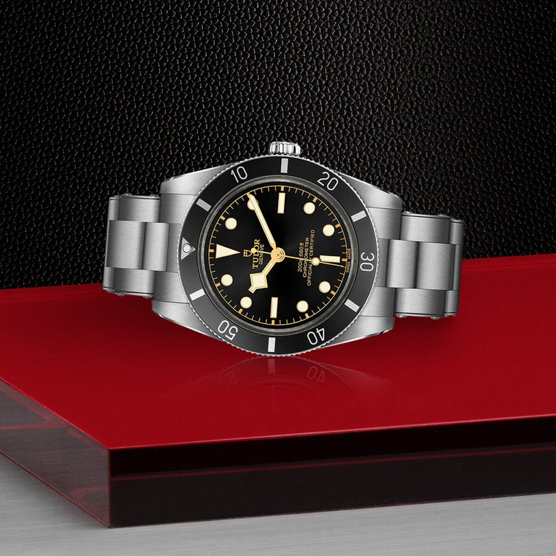 The TUDOR Black Bay 54 wristwatch with a black dial and bezel is displayed on a glossy red and black surface.