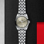 The TUDOR Black Bay One 31 unisex wristwatch, with a silver bracelet and champagne dial set with diamond hour markers, is displayed on a textured black surface beside a red accent.