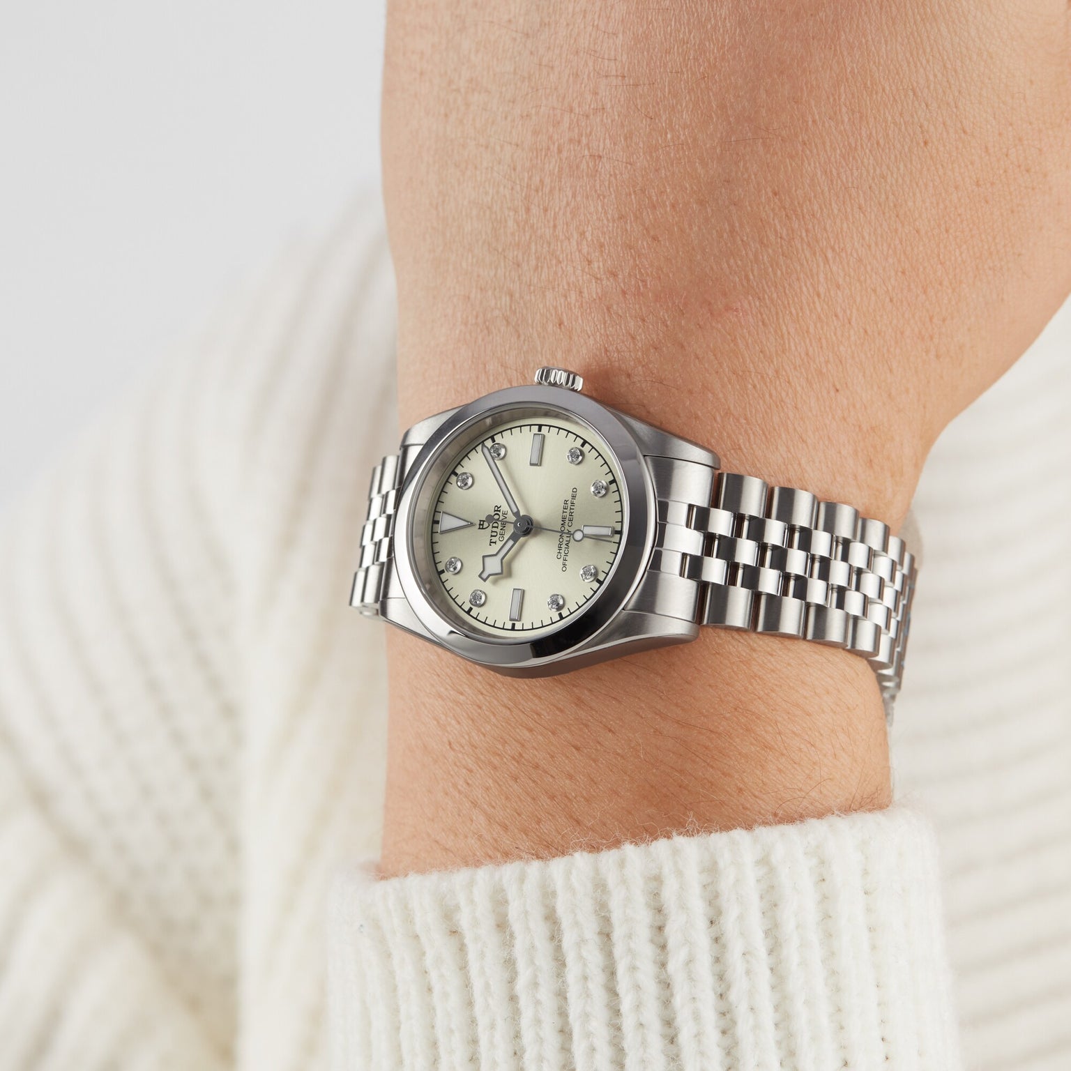 A TUDOR Black Bay One 31 wristwatch with a metal band on a persons wrist, paired with a white knit sweater.