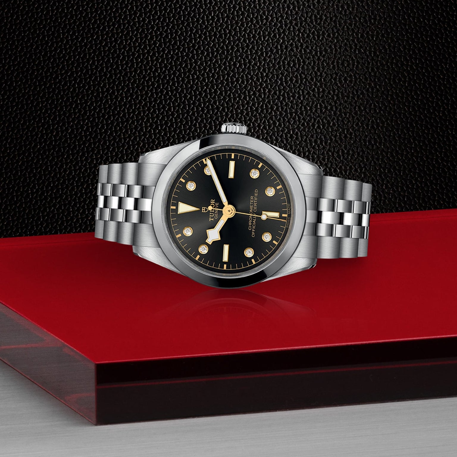 The TUDOR Black Bay One 36 unisex watch, with a stainless steel case, 316L steel bracelet, and black dial, is showcased on a red and black surface against a textured black background.