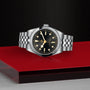 The TUDOR Black Bay One 36 unisex watch, with a stainless steel case, 316L steel bracelet, and black dial, is showcased on a red and black surface against a textured black background.
