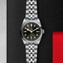 The TUDOR Black Bay One 36 unisex watch features a stainless steel case, black dial, gold hour markers, and a 316L steel bracelet, displayed on a textured black background with red and silver accents.