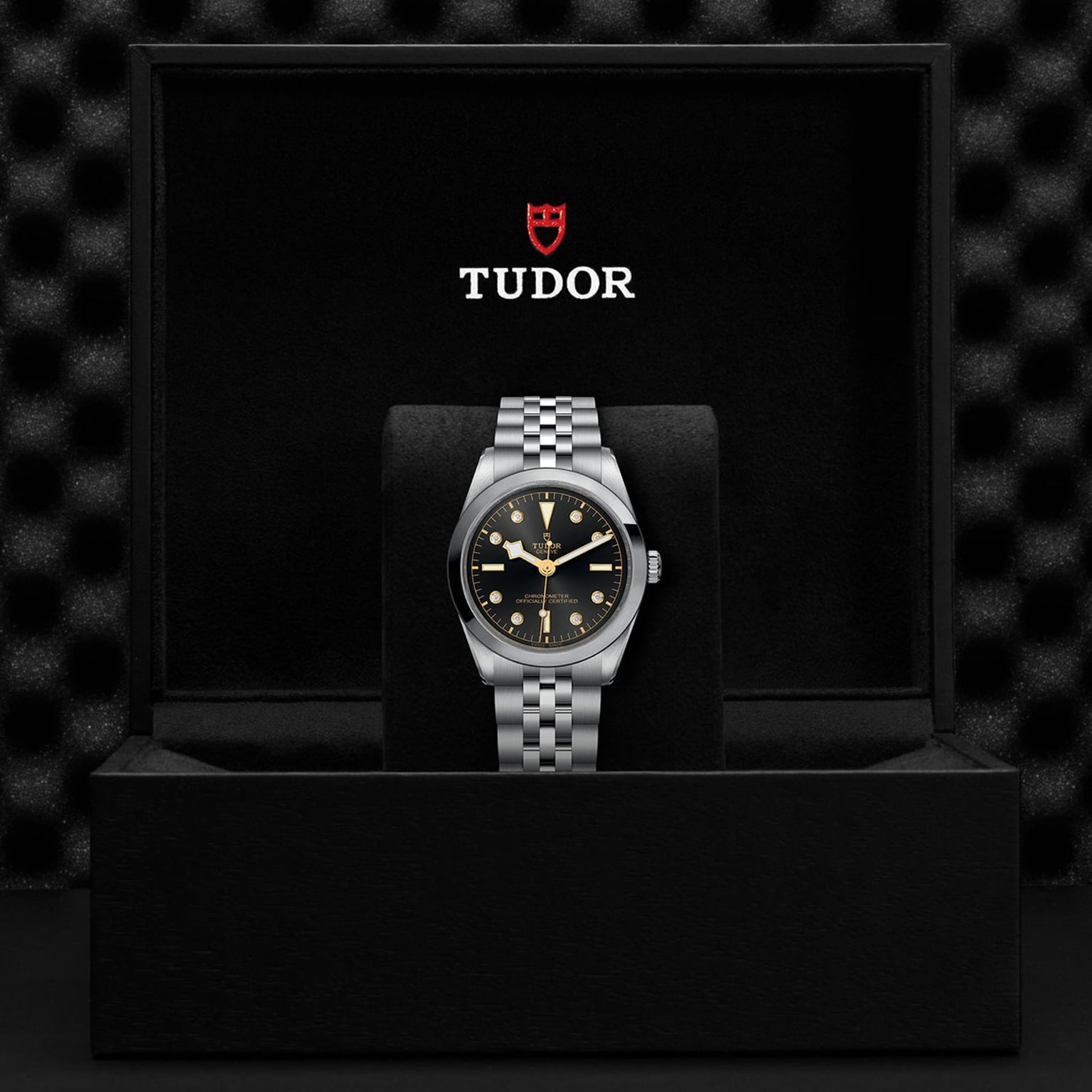 A TUDOR Black Bay One 36 unisex watch with a black dial and 316L steel bracelet is displayed in an open black TUDOR box, the logo clearly visible inside the lid.