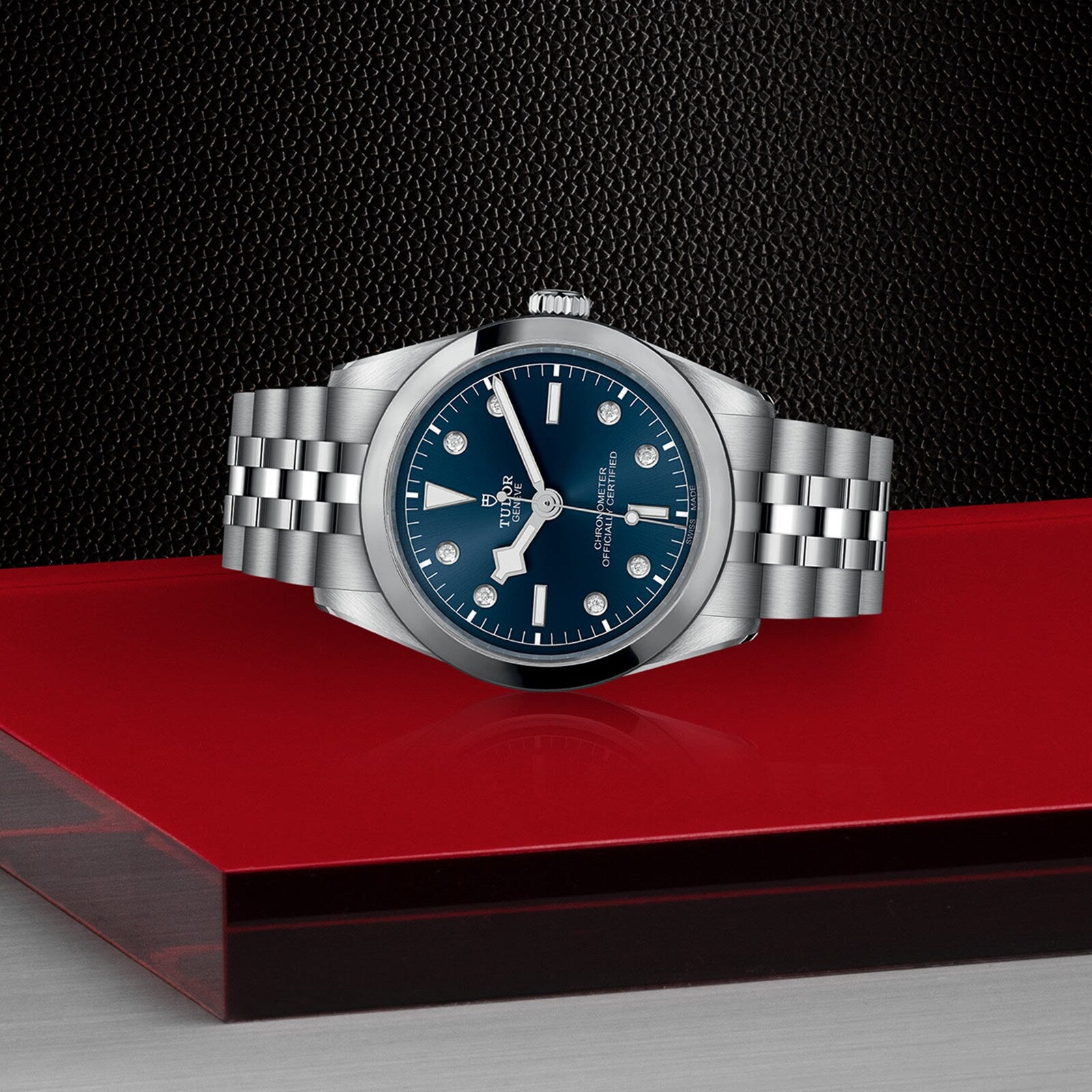 The TUDOR Black Bay One 36, a stainless steel unisex watch with a chic blue dial and metal bracelet inspired by Black Bay models, is displayed on a red and black geometric surface.