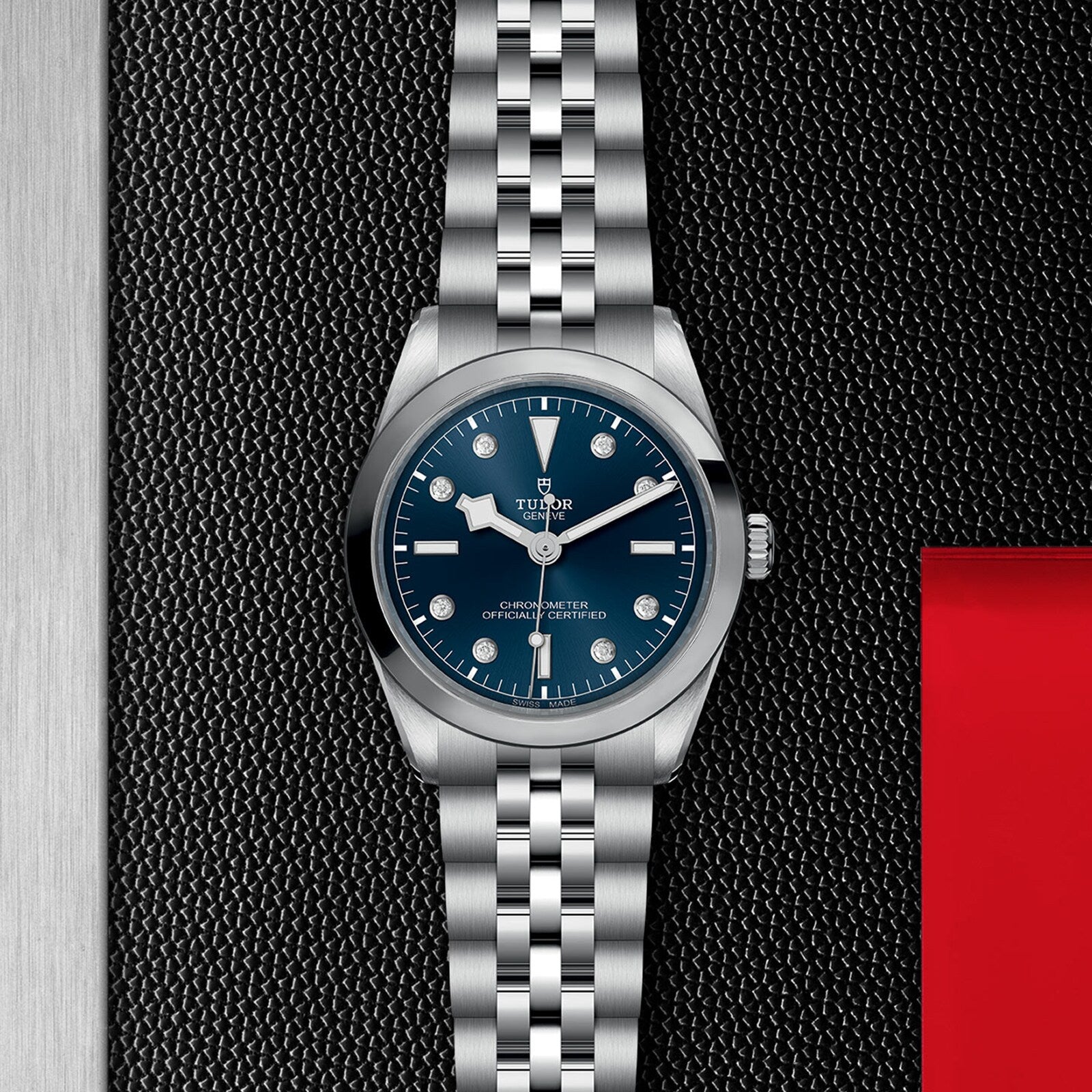 The TUDOR Black Bay One 36 wristwatch features a blue dial and silver metal bracelet, displayed against a textured black background.
