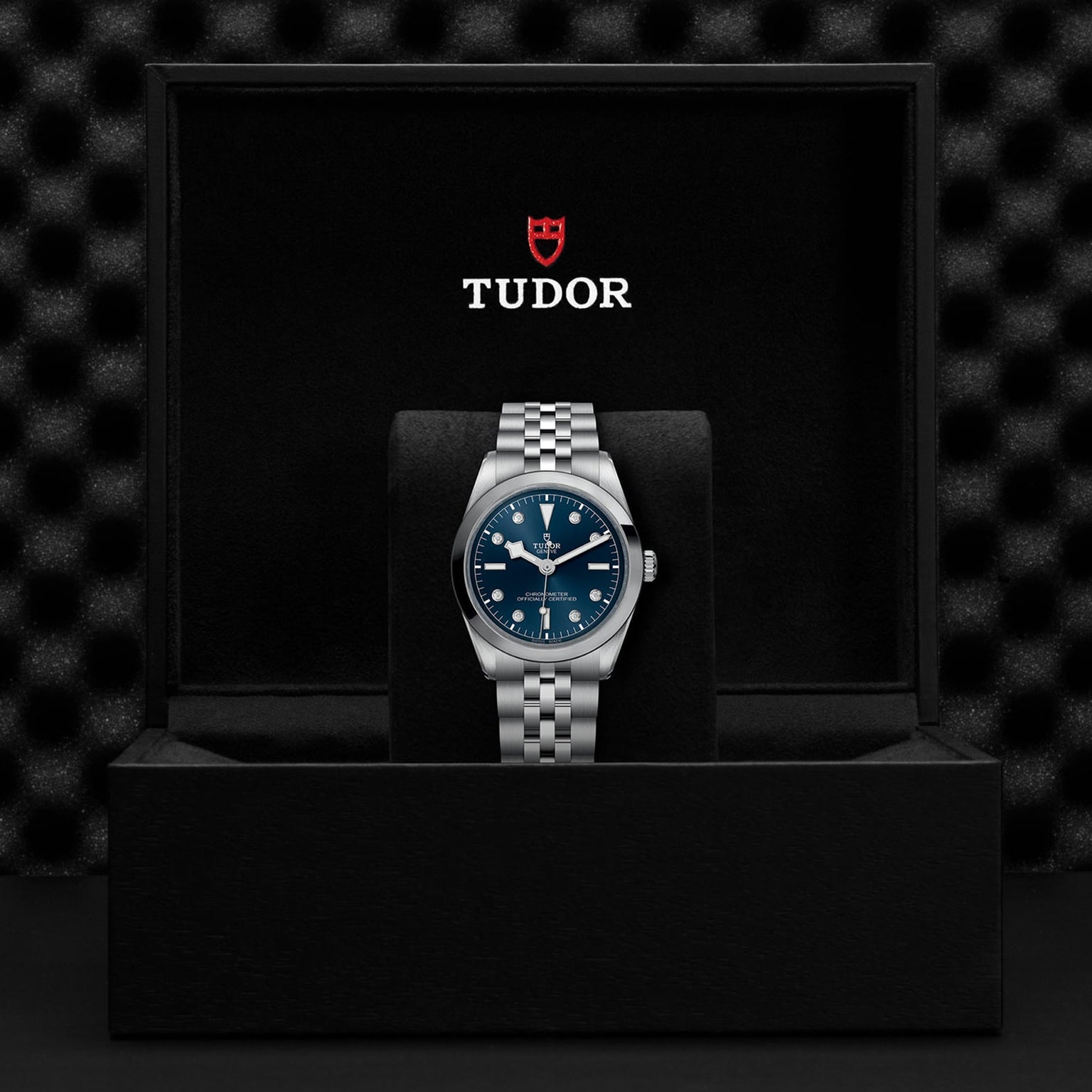 The TUDOR Black Bay One 36 wristwatch, featuring a blue dial, silver metal bracelet, and TUDOR rapid adjustment system, is displayed upright in an open black TUDOR-branded box.