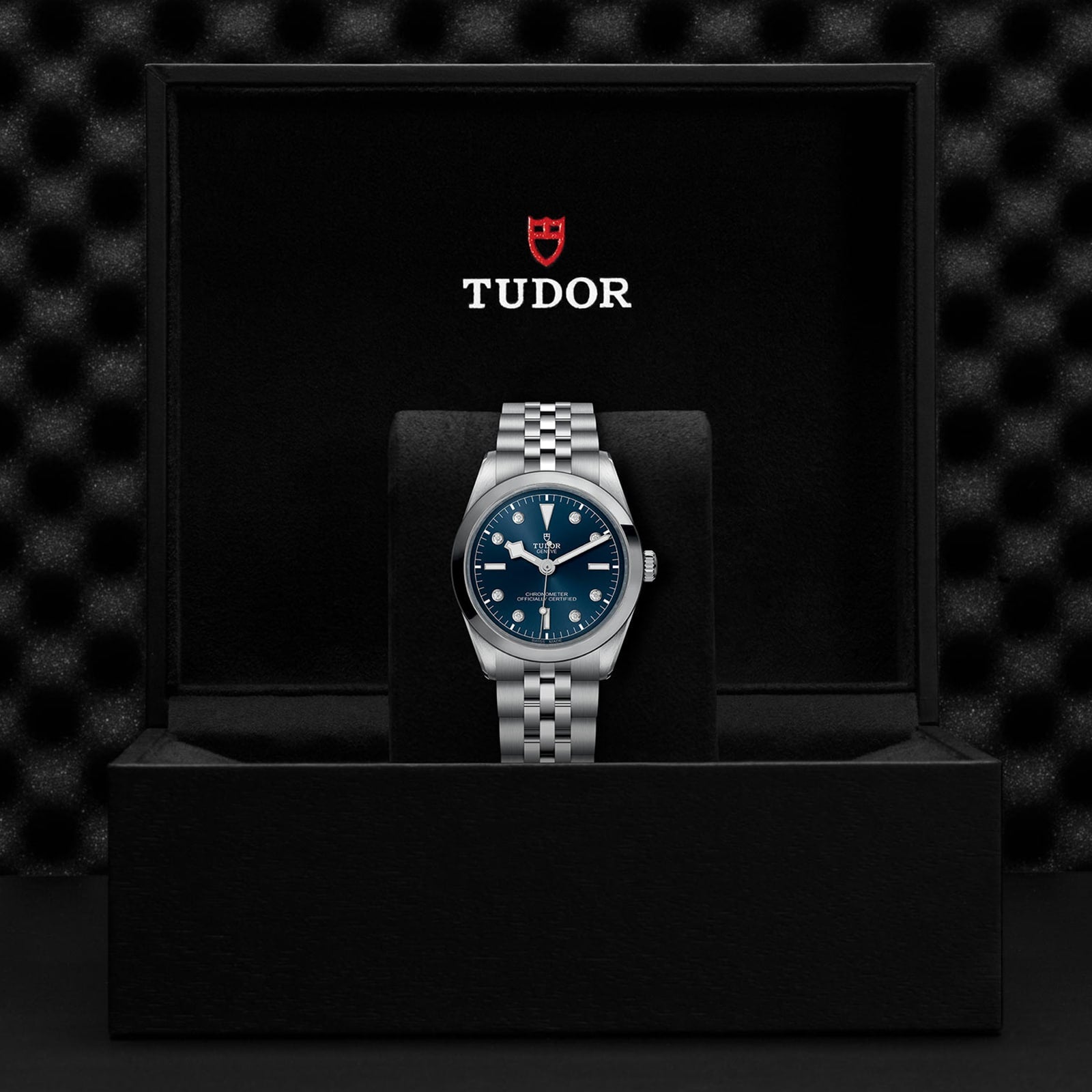 The TUDOR Black Bay One 36 wristwatch, featuring a blue dial, silver metal bracelet, and TUDOR rapid adjustment system, is displayed upright in an open black TUDOR-branded box.