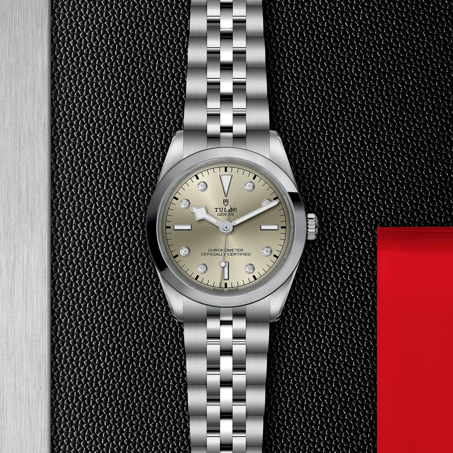 A TUDOR Black Bay One 36 wristwatch with a champagne dial, diamond hour markers, and a 316L steel bracelet is displayed on a textured black surface beside metal and red panels.