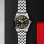 The TUDOR Black Bay One 39, a unisex wristwatch with a black dial, round hour markers, and a silver metal bracelet, is showcased on a textured black surface.