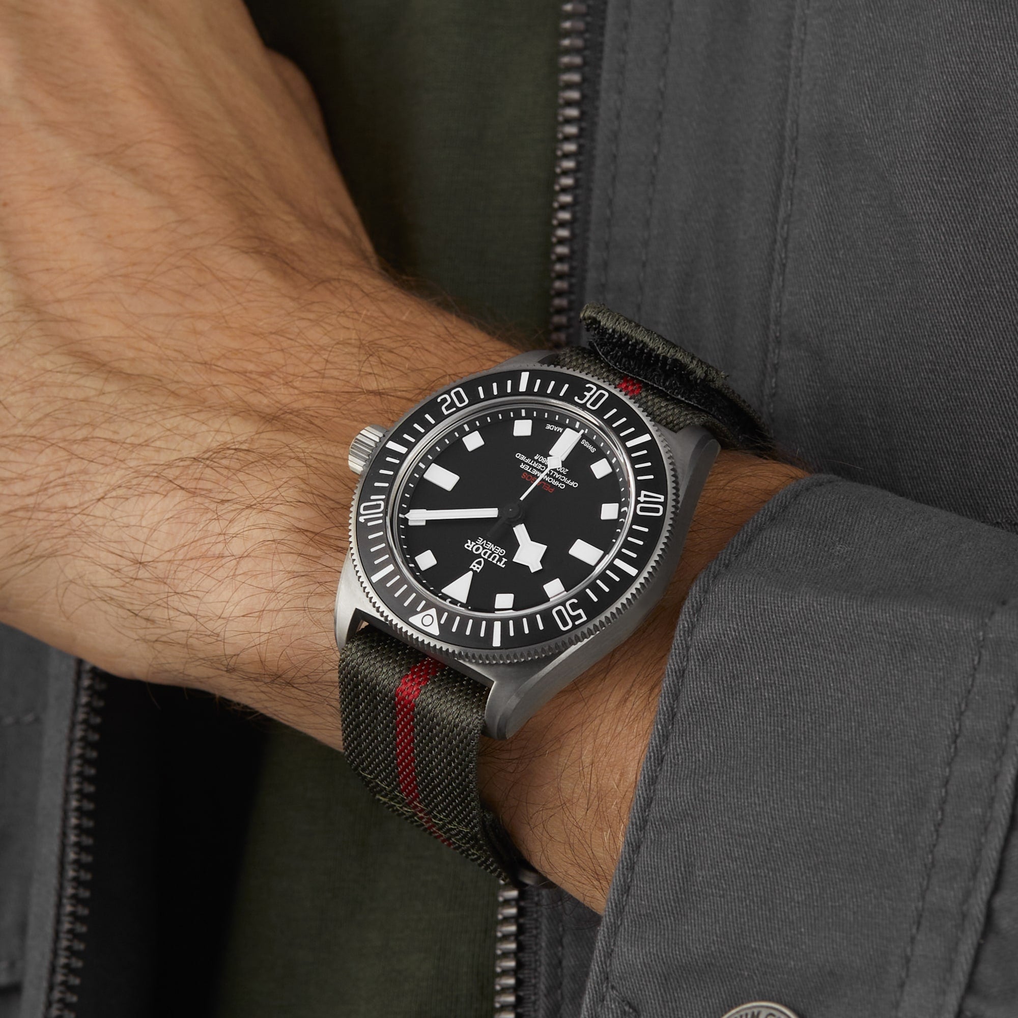 A person wearing a TUDOR Pelagos FXD diving watch with a black bezel and a green fabric strap accented by a red stripe.
