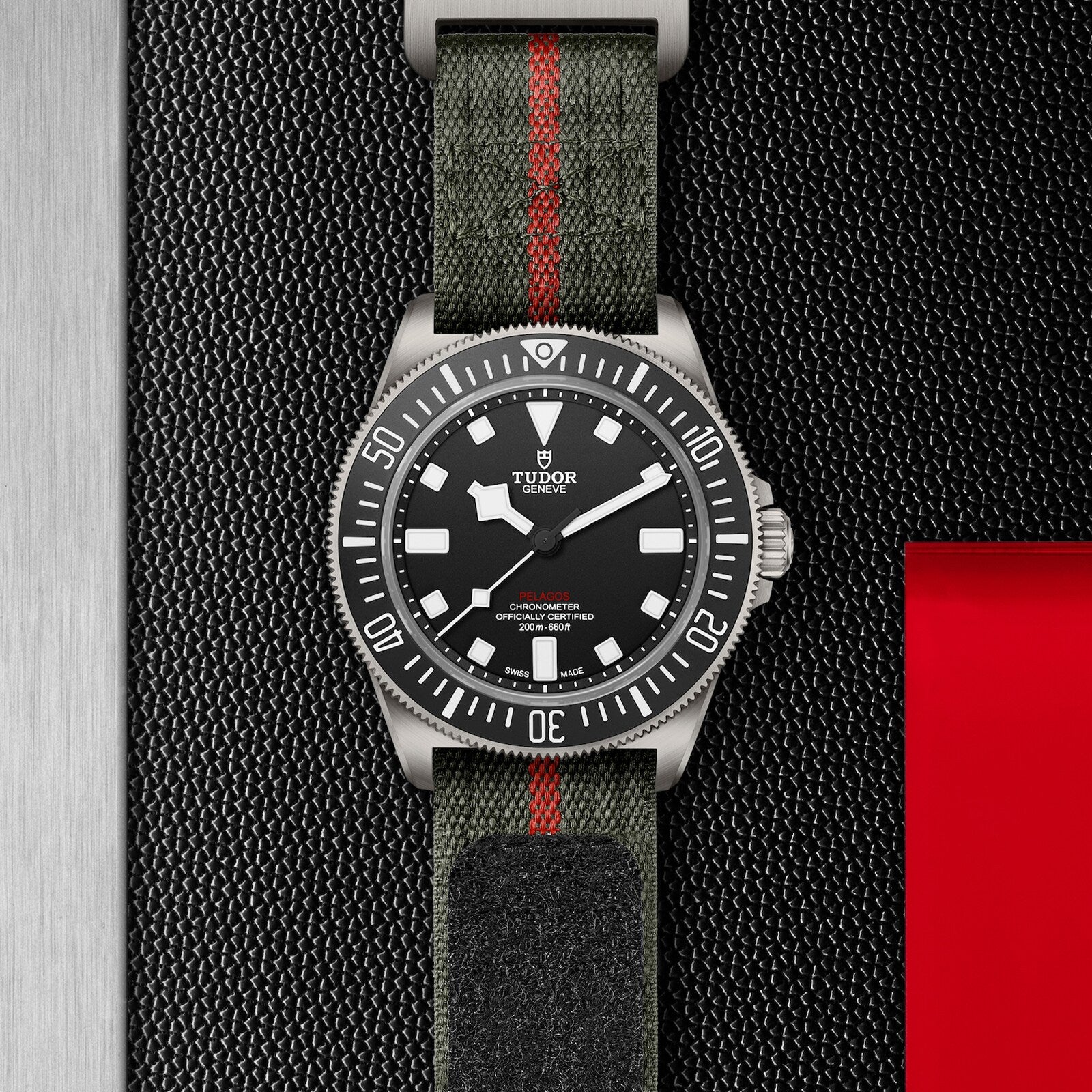 The TUDOR Pelagos FXD wristwatch features a black dial, white markers, and a green fabric strap with red stripe, set against a textured black and red backdrop—showcasing the heritage of TUDOR diving watches.