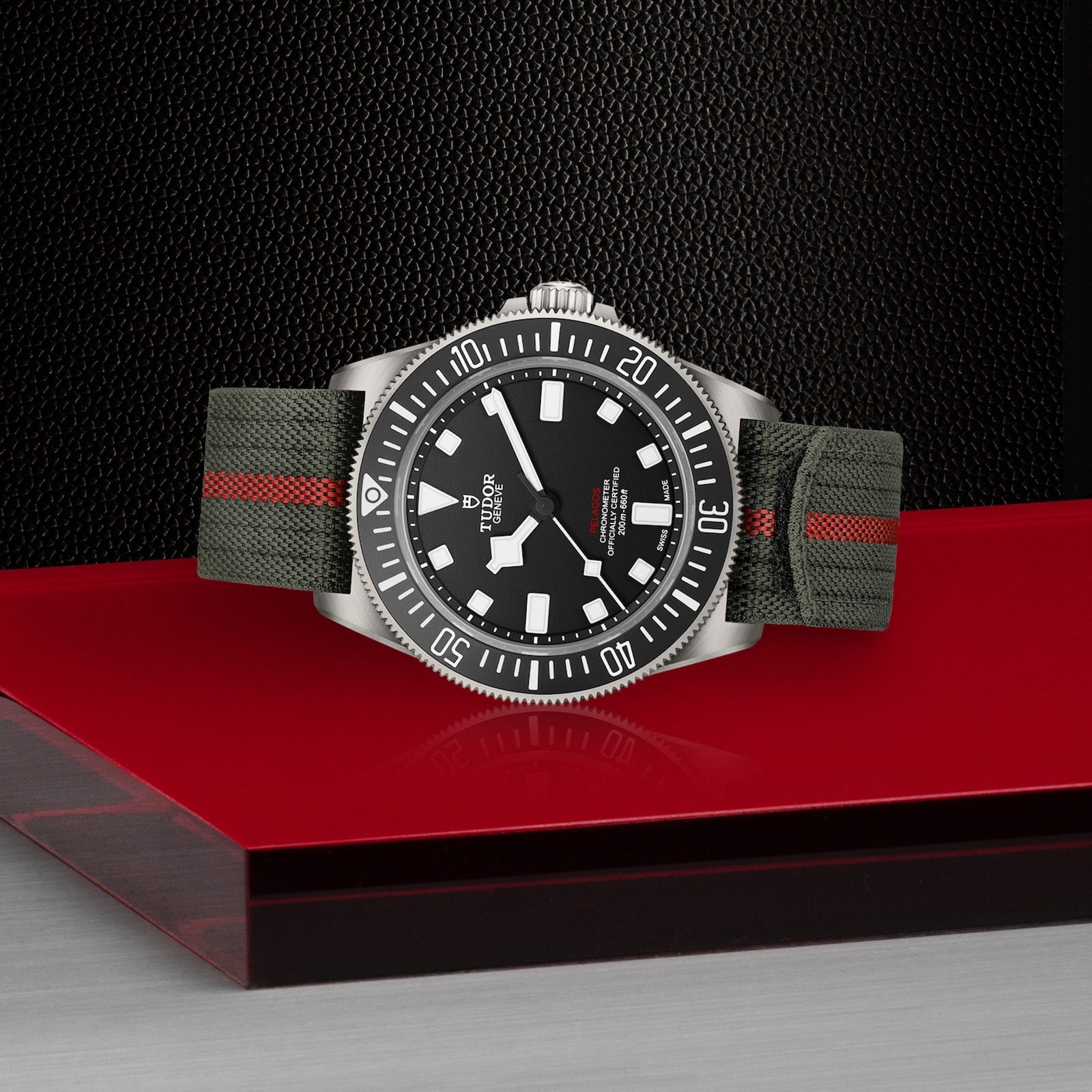 The TUDOR Pelagos FXD, featuring a black dial, white markers, rotating bezel, and green fabric strap with a red stripe, sits on a red and silver surface against a textured black background, evoking the Military Submariner spirit.