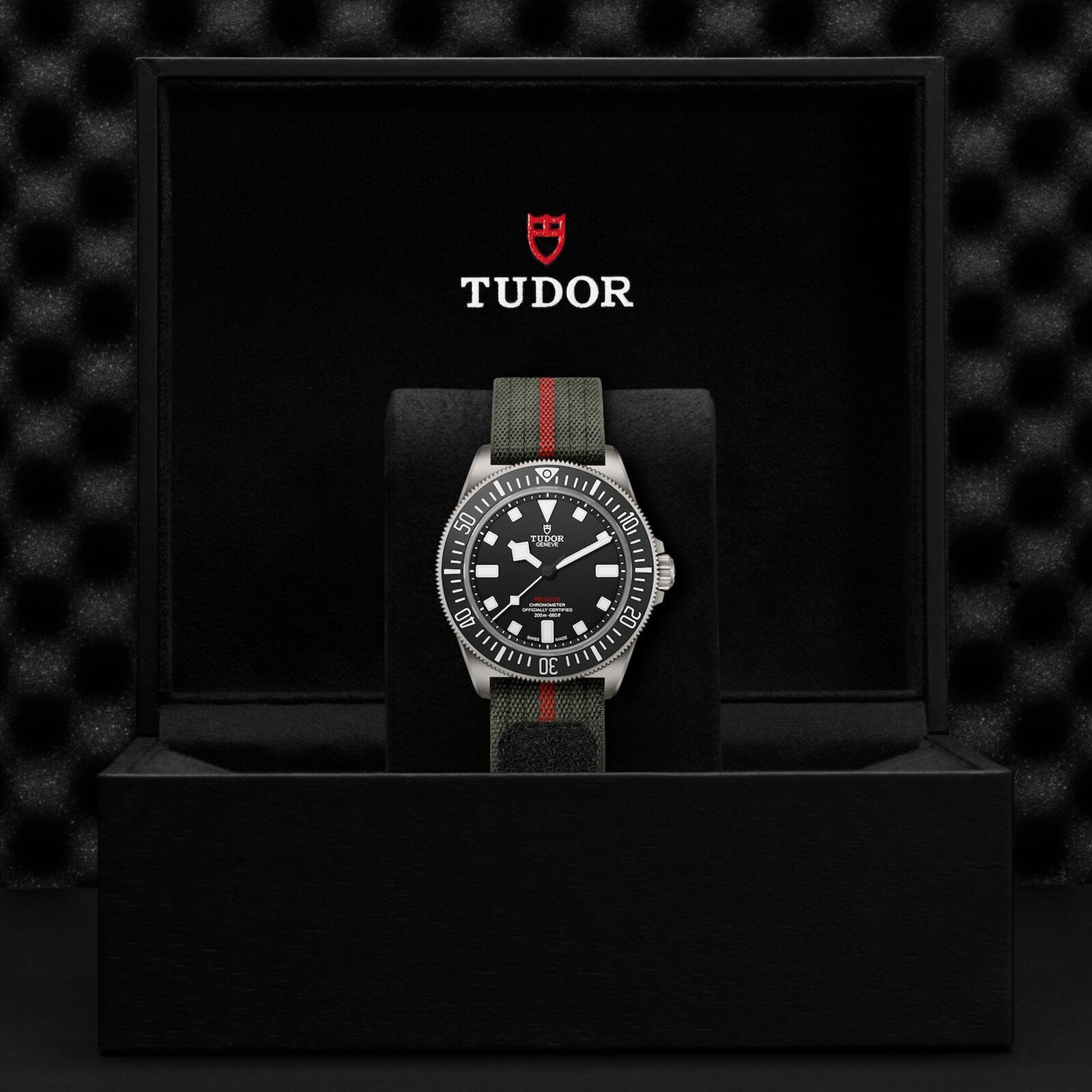 The TUDOR Pelagos FXD, inspired by Military Submariner heritage, features a black dial and striped fabric strap, displayed upright in a black presentation box with the TUDOR logo on the inner lid.