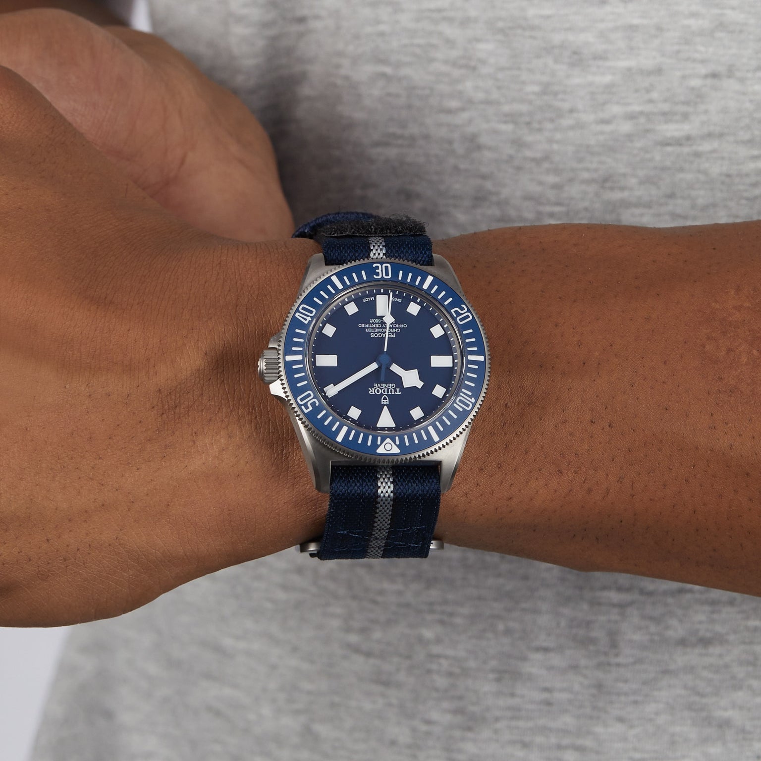 A person wears a TUDOR Pelagos FXD watch with a blue and silver dial and striped fabric strap on their left wrist, set against a gray shirt.