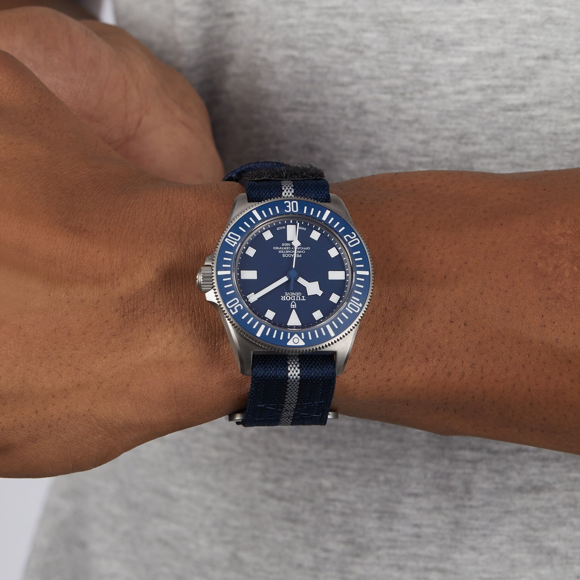 A person wears a TUDOR Pelagos FXD watch with a blue and silver dial and striped fabric strap on their left wrist, set against a gray shirt.