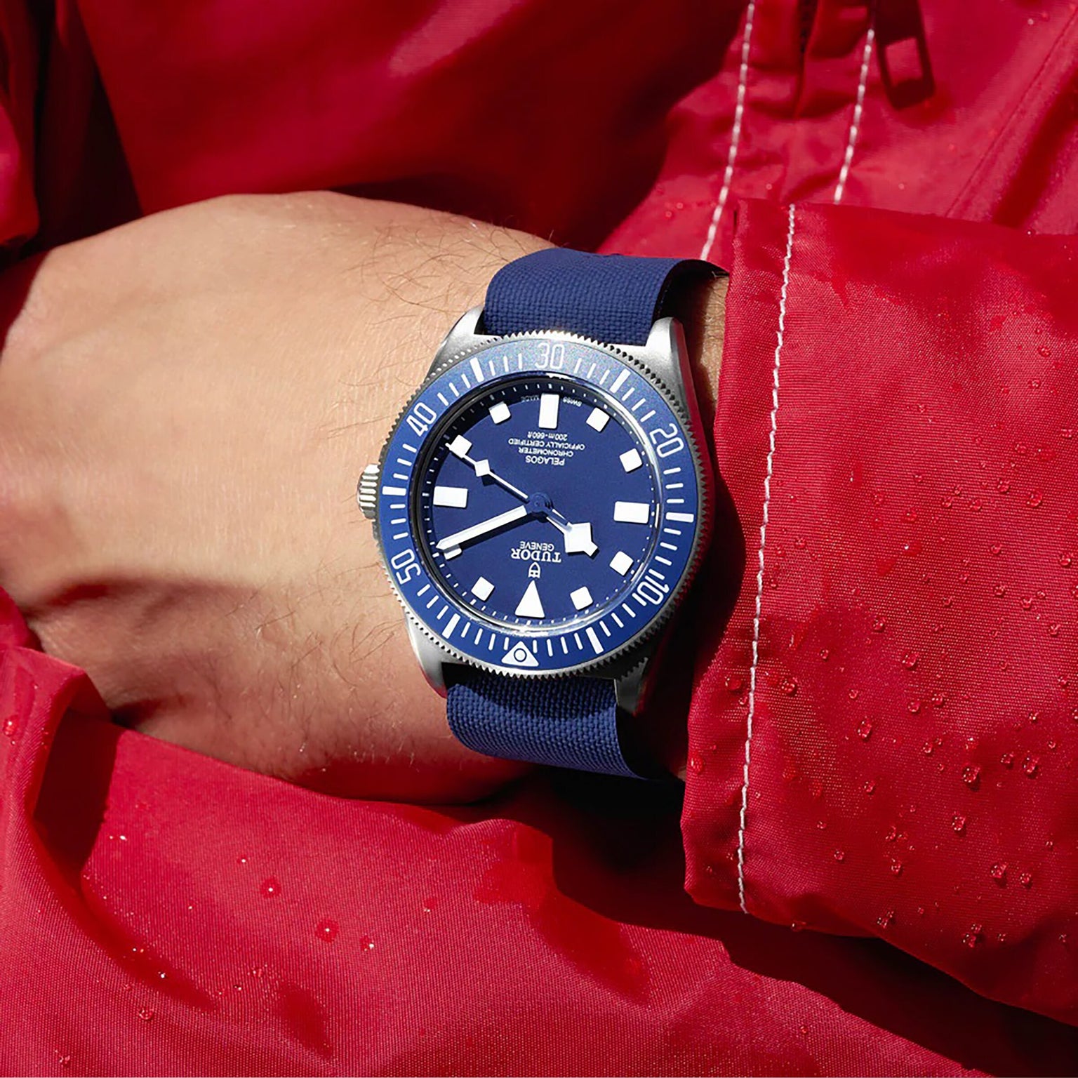 A person wearing the TUDOR Pelagos FXD, a blue diver’s watch with a navy strap, and a red jacket covered in water droplets.