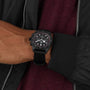 A person wearing a TUDOR Pelagos FXD Chrono wristwatch with a cycling-themed chronograph, dressed in a dark jacket over a maroon top.