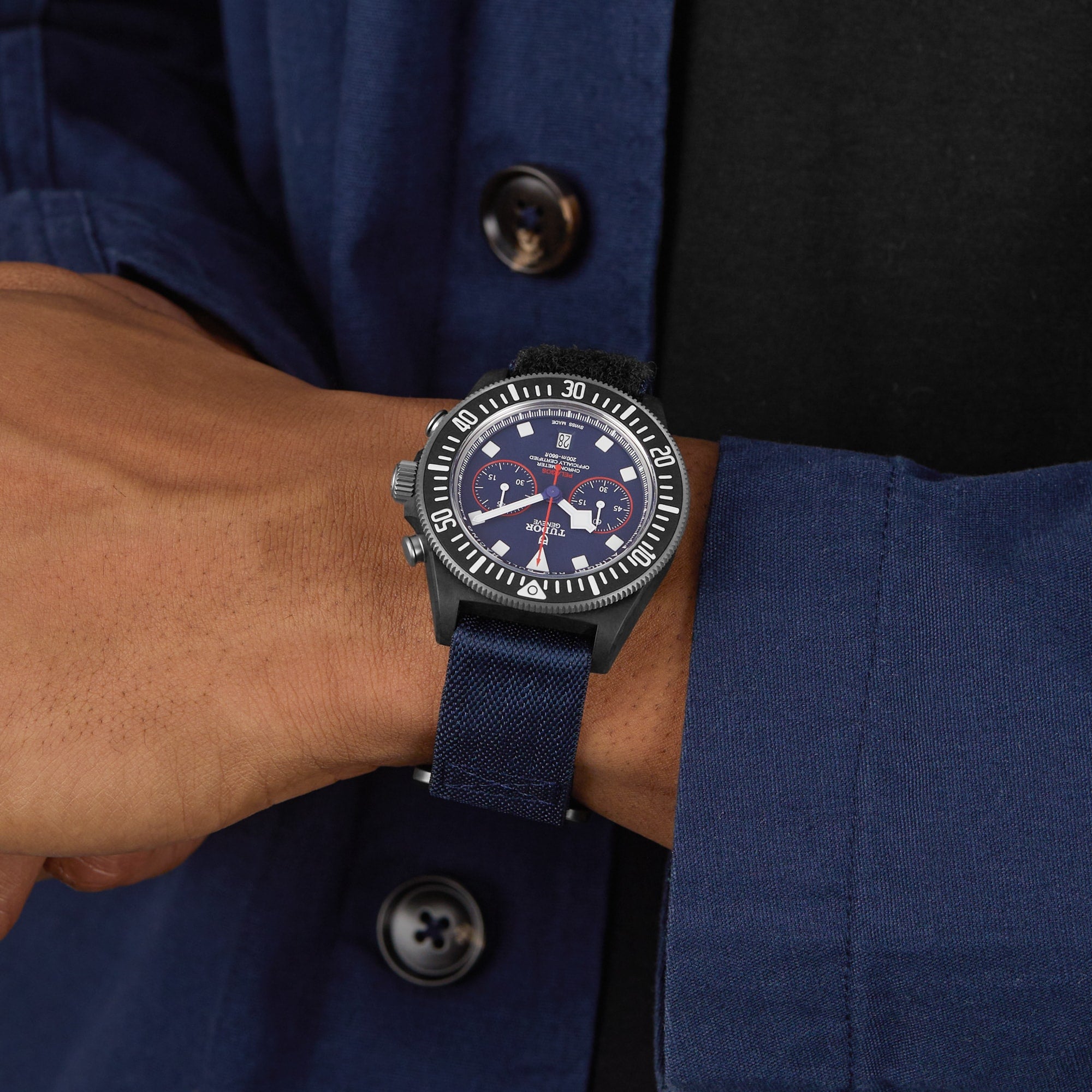 A person in a blue jacket showcases the TUDOR Pelagos FXD Chrono, a wristwatch with a blue strap and dark dial, crafted for underwater navigation and inspired by the French Navy.