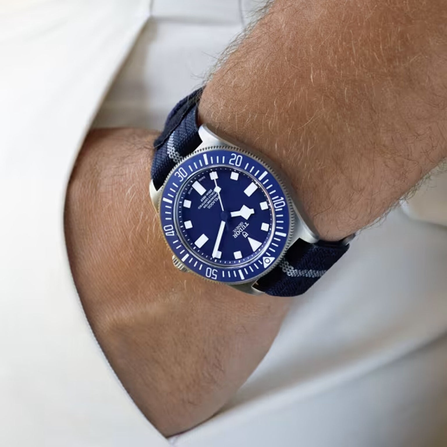 A person wears the TUDOR Pelagos FXD with a blue strap and titanium case, hand partly in pocket, dressed in light colors. This stylish watch is waterproof to 200m, offering both elegance and practical functionality.