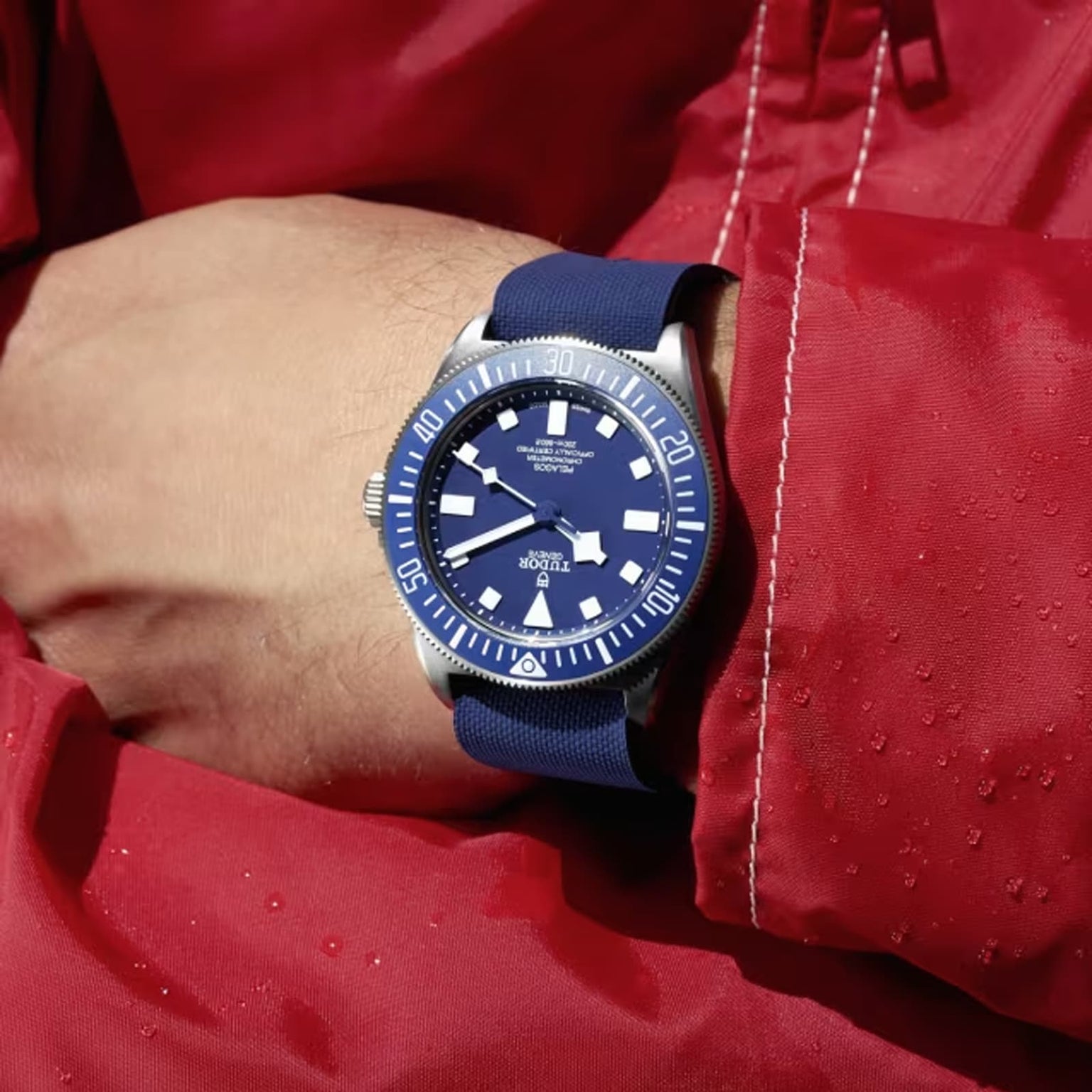A person in a red jacket with water droplets wears a TUDOR Pelagos FXD, featuring a titanium case and blue strap.
