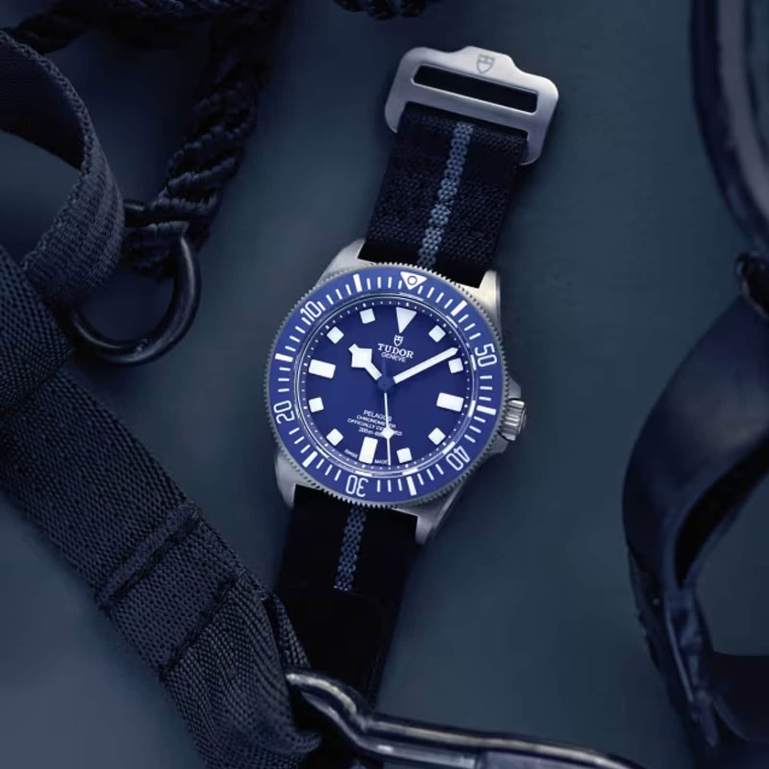 A TUDOR Pelagos FXD wristwatch with a blue face and bezel, white hour markers, and a titanium case is shown on a dark background, with black straps and buckles placed nearby.
