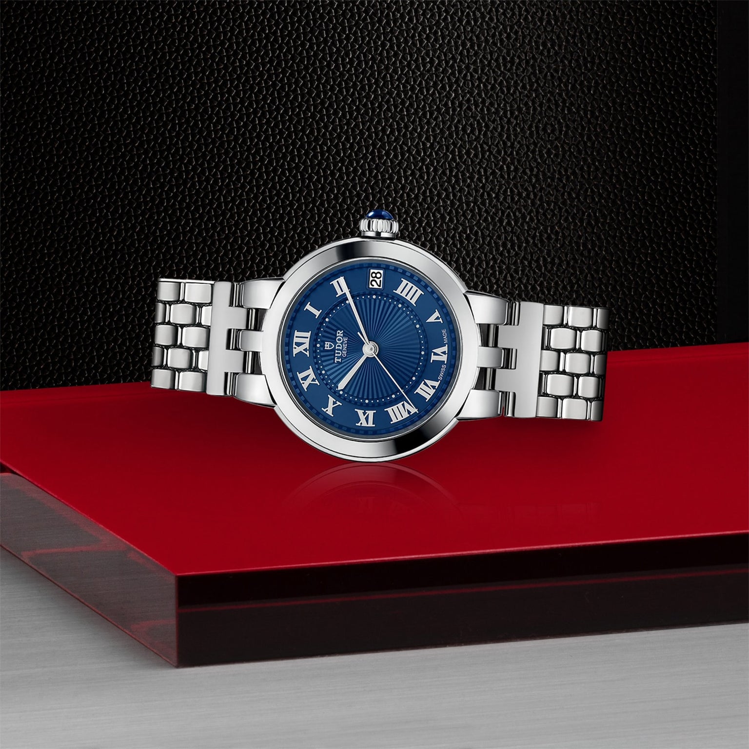The TUDOR Clair de Rose, a silver watch with a blue face and Roman numerals, is displayed on a red and black surface.
