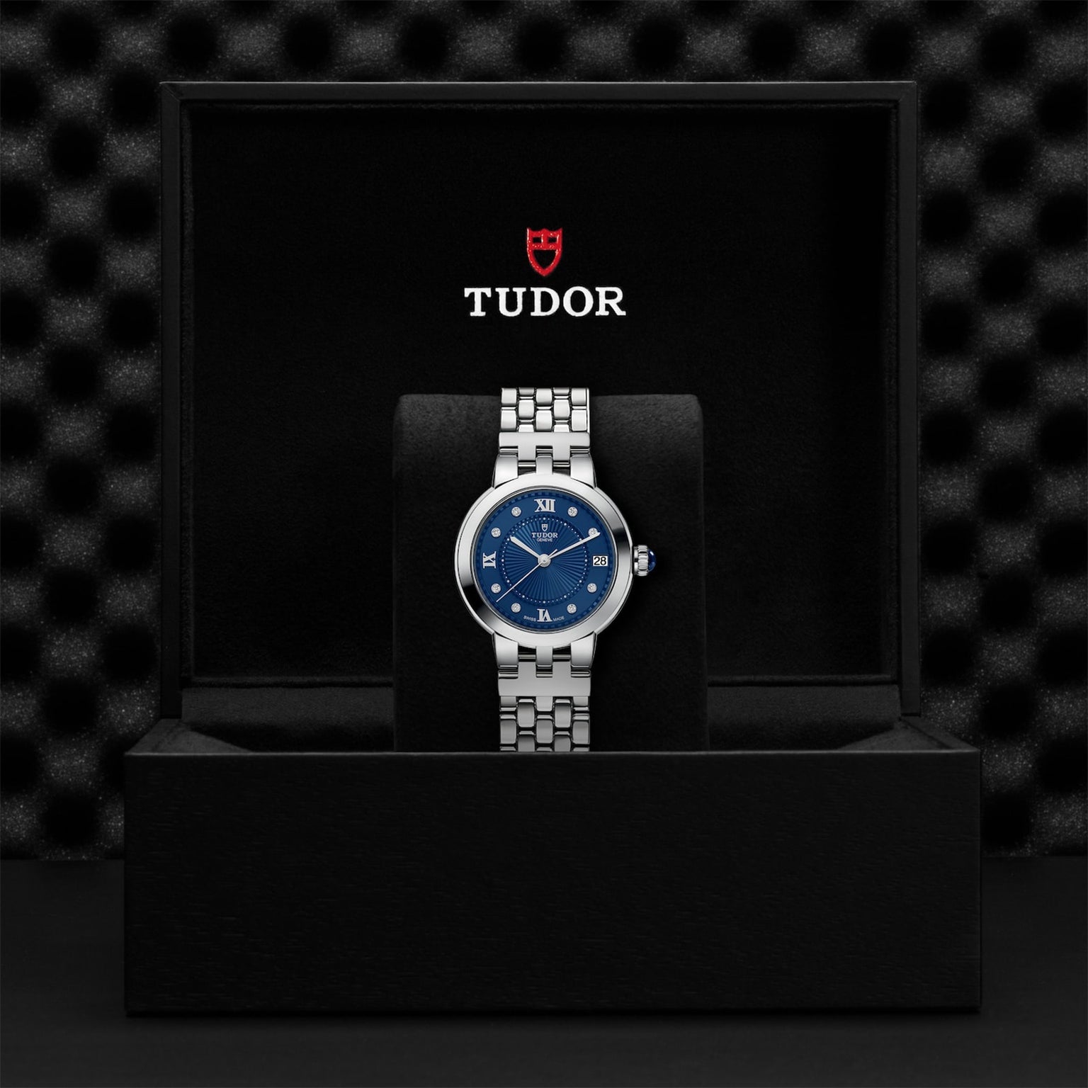 A silver TUDOR Clair de Rose wristwatch with a blue face, presented in a black TUDOR-branded display box.