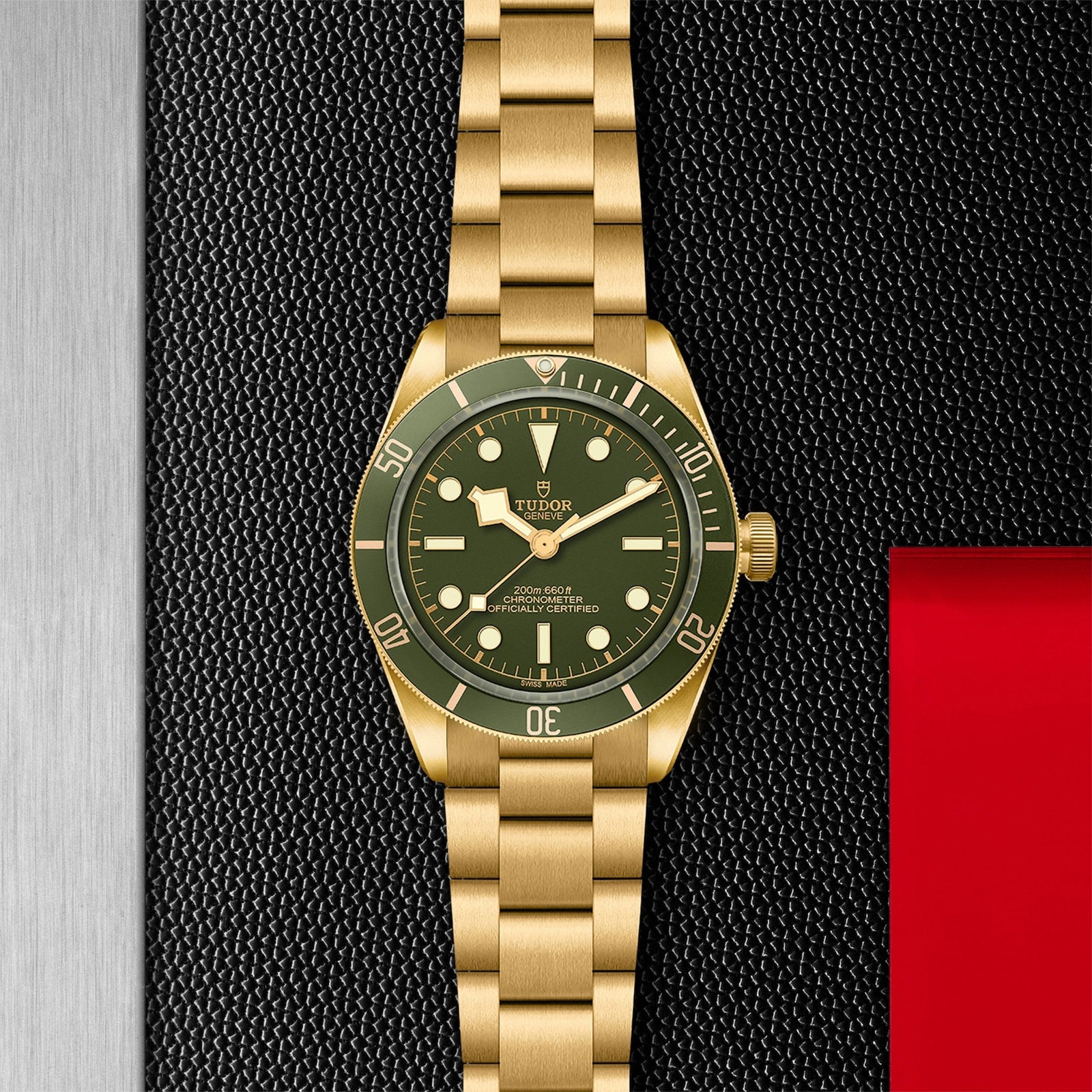 The TUDOR Black Bay 58 18K dive watch, featuring a gold case and green dial with bezel, is showcased on a textured black and silver background accented with red.