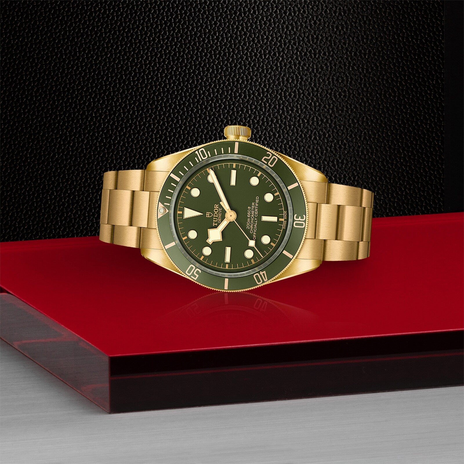 The TUDOR Black Bay 58 18K dive watch, featuring a gold case with green dial and bezel, is shown on a reflective red and black surface against a textured black background, highlighting its signature style.