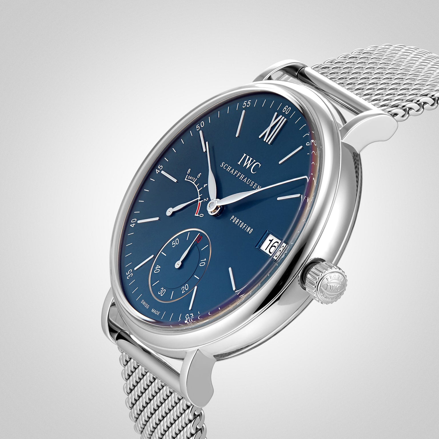 The Portofino Hand-Wound Eight Days by IWC Schaffhausen features a blue dial, mesh band, date display at 3 oclock, and a sub-dial at 6 oclock, displayed against a light grey background.