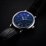 The IWC Schaffhausen Portofino Automatic features a blue dial, silver hands and markers, a date window at 3 o’clock, and a black leather strap, exemplifying Swiss precision on a dark surface.