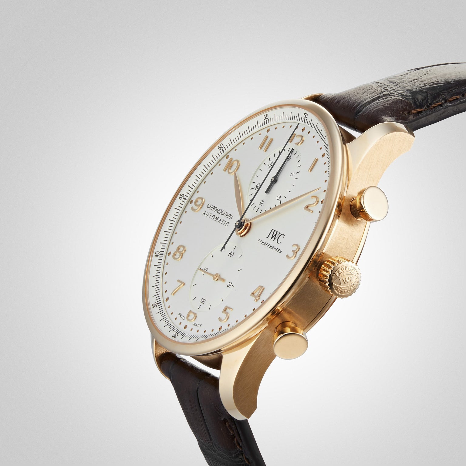 The IWC Schaffhausen Portugieser Chronograph, with a gold case, white dial, black and gold numerals, two subdials, and a brown leather strap, is elegantly displayed at an angle on a light background.