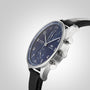 A close-up shows the IWC Schaffhausen Portugieser Chronograph with a blue dial, stainless steel case, black leather strap, and two side pushers set against a plain gray background.
