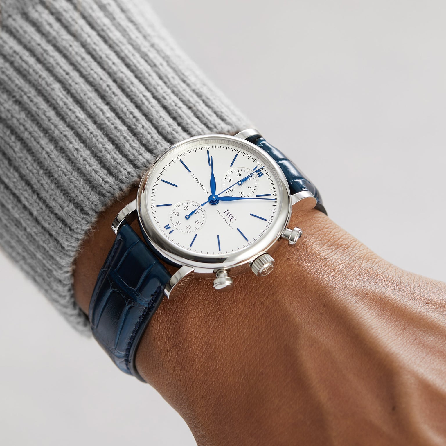 A person wearing the IWC Schaffhausen Portofino Chronograph, featuring a silver stainless steel case, white dial with blue markers, and a blue alligator leather strap.